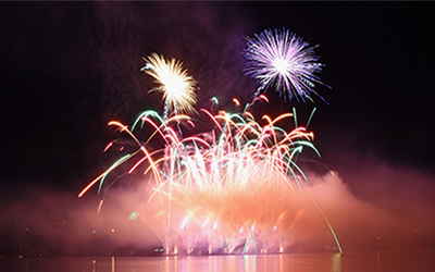 L’image montre un magnifique spectacle pyrotechnique au-dessus d’un plan d’eau. Plusieurs feux d’artifice multicolores explosent simultanément, créant un affichage spectaculaire et captivant. La fumée et les effets lumineux des feux d’artifice se reflètent sur la surface de l’eau. L’arrière-plan est sombre, permettant aux couleurs vives des feux d’artifice de se démarquer. L’atmosphère générale de l’image est animée et festive, typique d’un spectacle de feux d’artifice.