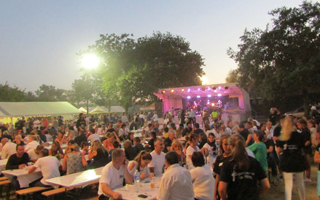Cette image montre la scène animée et festive du grand restaurant, lors de Gujan-Mestras en Fêtes en plein air. La photo est prise en soirée, à la tombée de la nuit, comme l'indique la lumière douce et le ciel crépusculaire. Une grande foule de personnes est rassemblée autour de tables disposées sur une pelouse, partageant un repas et profitant de l'ambiance. Au fond, une scène est installée, avec un groupe de musique.