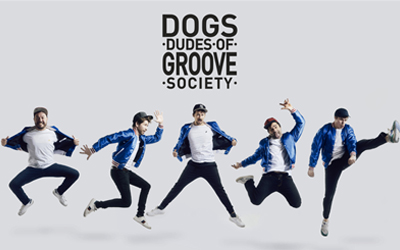 L'image montre le groupe Dudes of Groove Society (DOGS) en pleine performance sur scène. Les membres du groupe, vêtus de tenues aux couleurs vives, sont capturés dans une pose dynamique, reflétant l'énergie et la convivialité de leurs prestations. Le fond est blanc, mettant en avant les musiciens et leur instrumentation, tandis que des jeux de lumière ajoutent à l'ambiance festive.