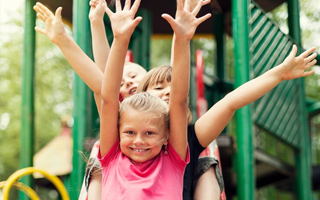Cette image représente trois enfants, deux filles et un garçon, qui descendent joyeusement un toboggan dans une aire de jeux en plein air. Leurs bras sont levés en l'air, exprimant une joie et une excitation palpable. La scène est baignée d'une lumière naturelle et chaleureuse, suggérant une journée ensoleillée. Les couleurs vives des vêtements des enfants et du toboggan contribuent à l'atmosphère joyeuse et dynamique de l'image.
