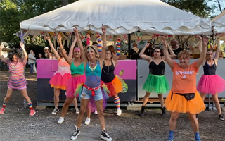 L'image montre un groupe de femmes en costumes colorés, ressemblant à des tutus, qui dansent en groupe devant une tente blanche. Elles ont les bras levés et semblent s'amuser. Les costumes varient en couleur, y compris le rose, l'orange, le vert et le bleu.