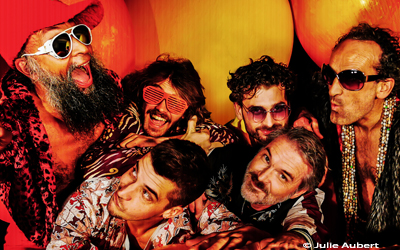 L'image représente le groupe Tha Dunciz, surnommé "Les cancres du funk", dans une ambiance colorée et décalée, fidèle à leur univers musical. Sur scène, les six membres du groupe sont vêtus de tenues vives et stylisées, avec des couleurs éclatantes comme le rouge, le jaune et le violet. Le chanteur, Zonk, adopte une posture dynamique, capturant l'attention du public avec son énergie communicative.